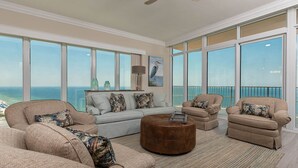 A spacious living room with a large window overlooking the ocean, a gray sofa, two armchairs, and a brown ottoman. - Phoenix Gulf Tower 2604 Seaside Escape (Orange Beach)