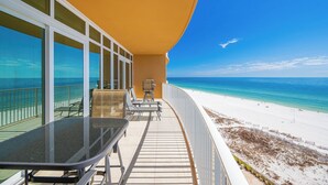 Ein Balkon mit Glastisch und Stühlen, der Blick auf einen Strand mit klarem, blauem Wasser und sandigem Ufer bietet.