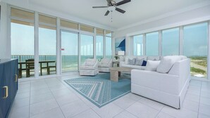 Un vaste espace de vie comprenant un canapé blanc, deux fauteuils et une table basse. De grandes fenêtres offrent une vue sur l’océan et donnent accès à un balcon.