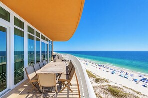 Un balcon avec une table et des chaises en bois, donnant sur une plage parsemée de parasols et où se trouvent des gens.