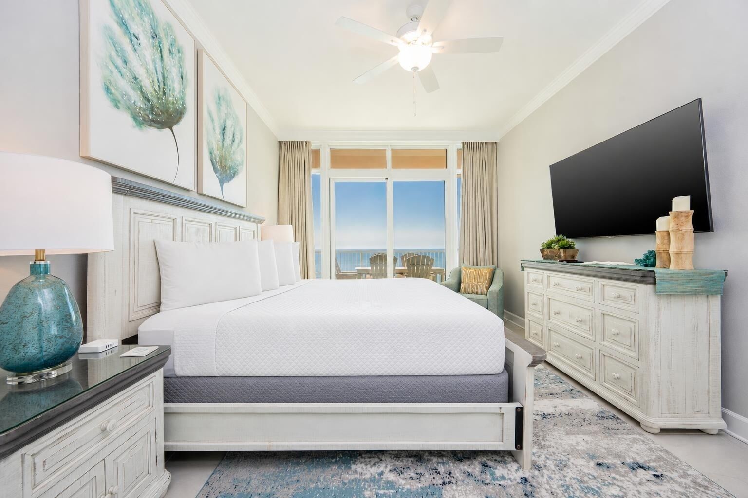 A bedroom with a bed, a TV, a ceiling fan, a dresser, and a view of the ocean.