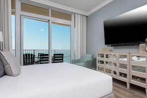A hotel room with a large bed, a TV, a dresser, and a balcony with chairs.