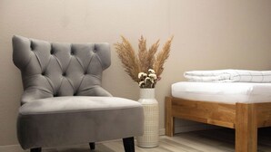 A modern bedroom with a grey upholstered armchair, a wooden bed frame, a white bedside table, and a vase with dried flowers. - Kuzey Apartments (Kappel-Grafenhausen)