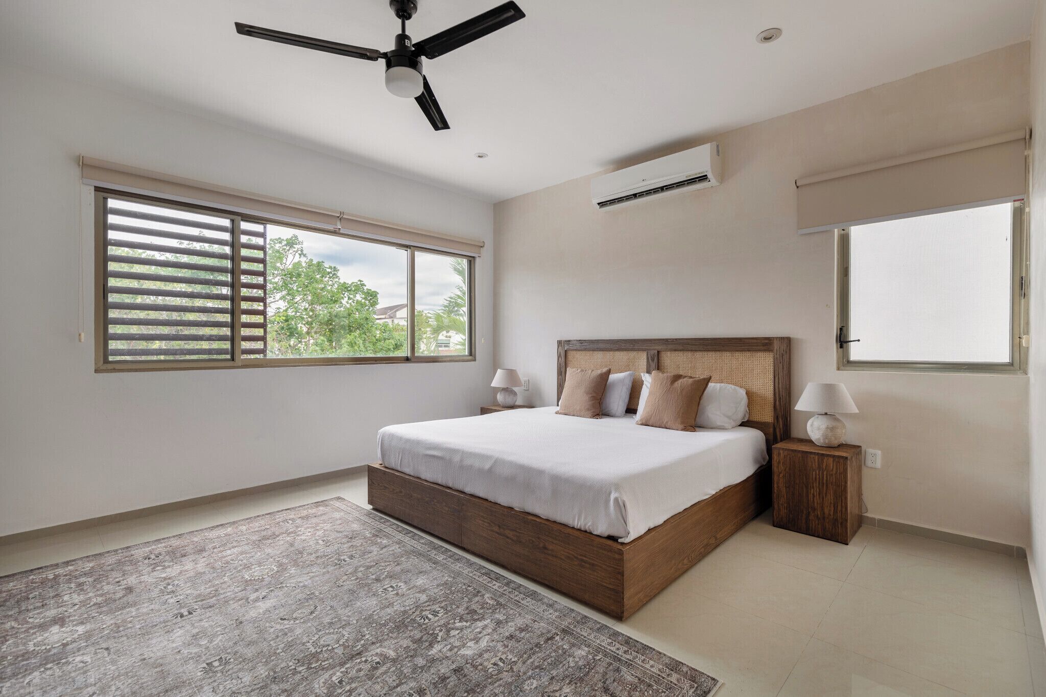 A bedroom with a large bed, a ceiling fan, an air conditioner, and a view of greenery outside.