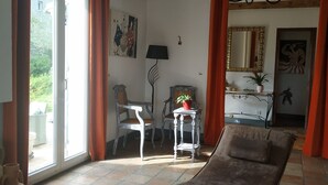 Una sala de estar acogedora con un sofá, una mesa con lámpara y un sillón. Se aprecia una ventana con cortinas, un cuadro en la pared y una araña.