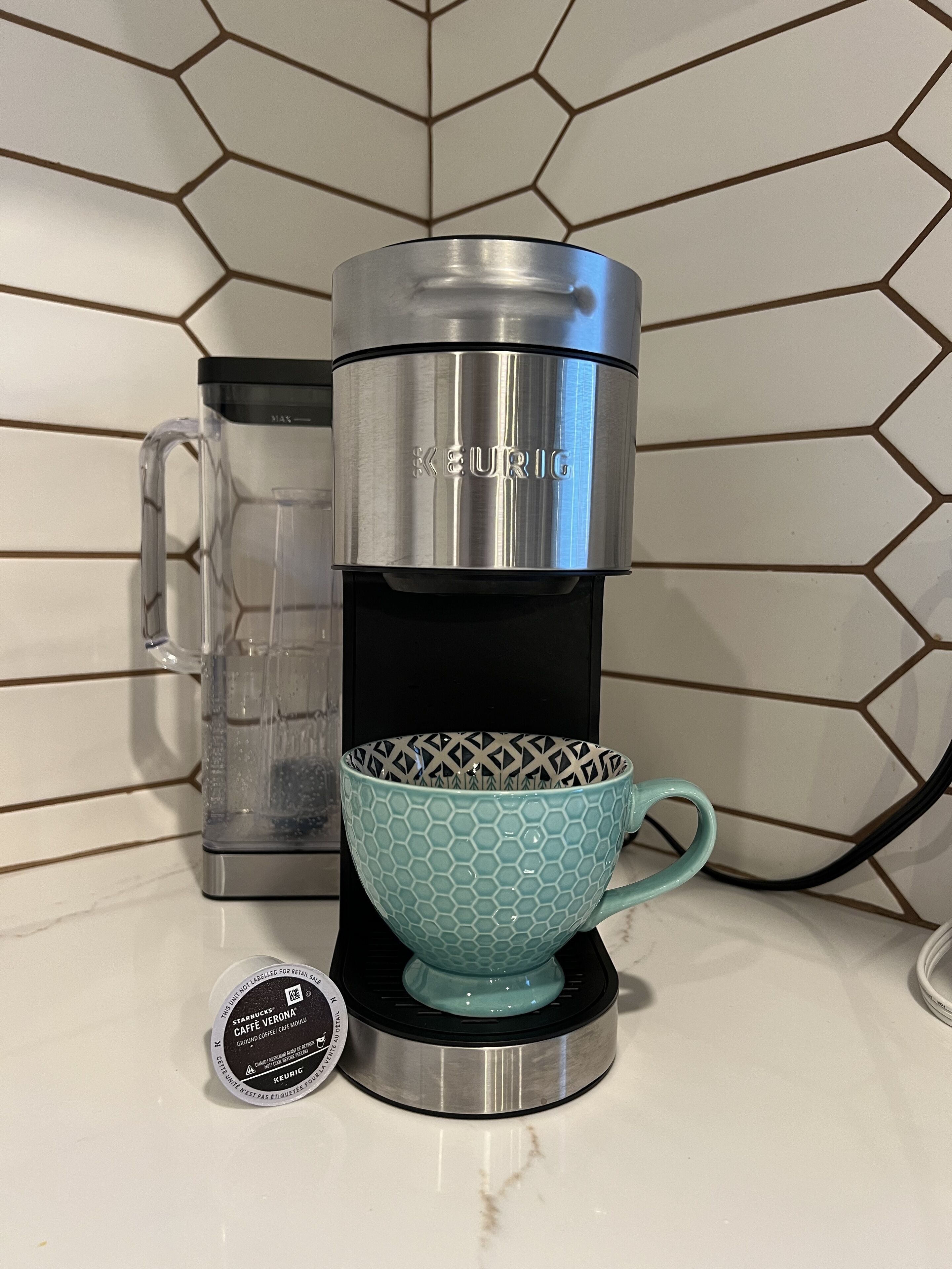 A Keurig coffee maker with a clear mug and a coffee pod on a white tiled backsplash.