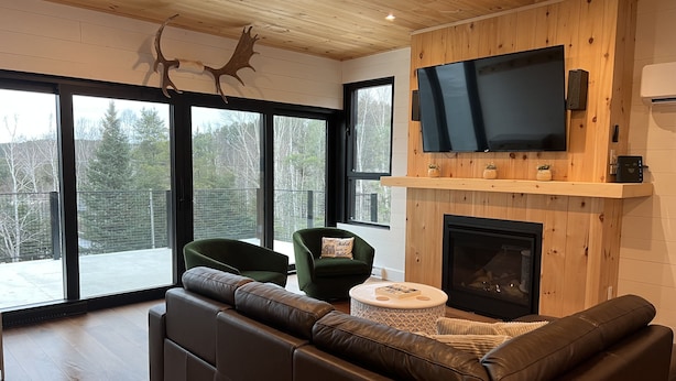 A modern living room with a leather sofa, two green armchairs, a fireplace, and a large TV mounted on the wall. - New construction, This Chalet is ready to enjoy all seasons ! (Saint-Michel-Des-Saints)