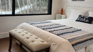 A bedroom with a large window, a bed with a white comforter, a bench, and a painting on the wall. - New construction, This Chalet is ready to enjoy all seasons ! (Saint-Michel-Des-Saints)