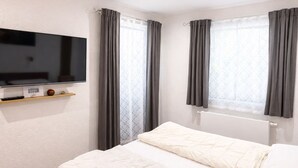 A bedroom with a bed, a TV mounted on the wall, and a window with curtains. - Ferienhaus Grundmann - im Allgäu mit Wunderschönem Berg- Seepanorama (Lechbruck am See)