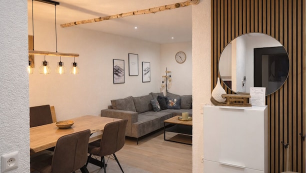 A modern living room with a wooden dining table, grey sofa, and a white cabinet with a mirror. - Ferienhaus Grundmann - im Allgäu mit Wunderschönem Berg- Seepanorama (Lechbruck am See)