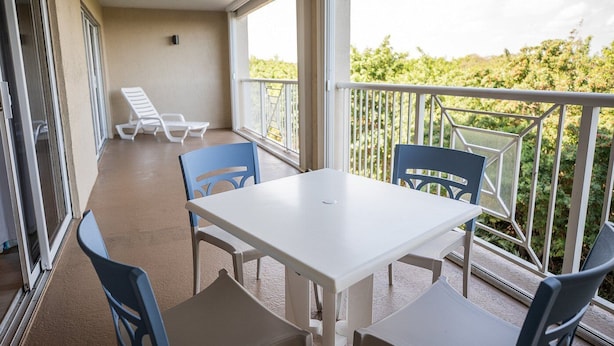 Uma varanda com mesa branca e cadeiras azuis, uma cadeira branca, e vista para o verde.