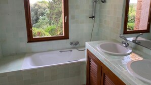 Une salle de bain avec une baignoire, deux lavabos, une douche et une fenêtre donnant sur un espace verdoyant.