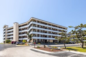 A multi-story hotel with a modern design, featuring balconies on each floor and a parking area in front.