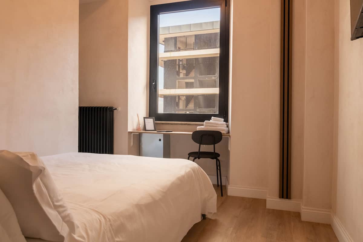 Quarto com cama, escrivaninha com cadeira, radiador e janela com vista para edifícios.