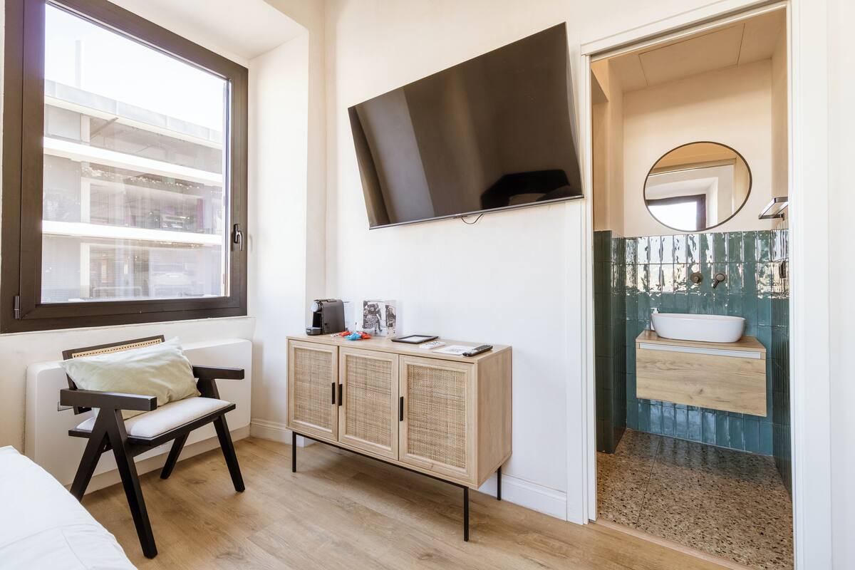 Quarto com cama, janela, televisão, armário e banheiro com pia e espelho.