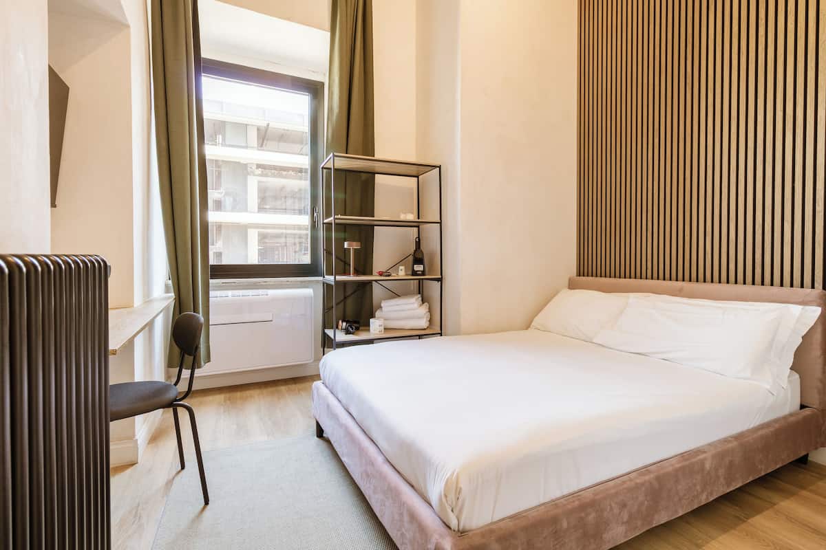 Quarto de hotel com cama, uma cadeira, radiador, janela com cortinas, prateleira com toalhas e painel de parede de madeira.