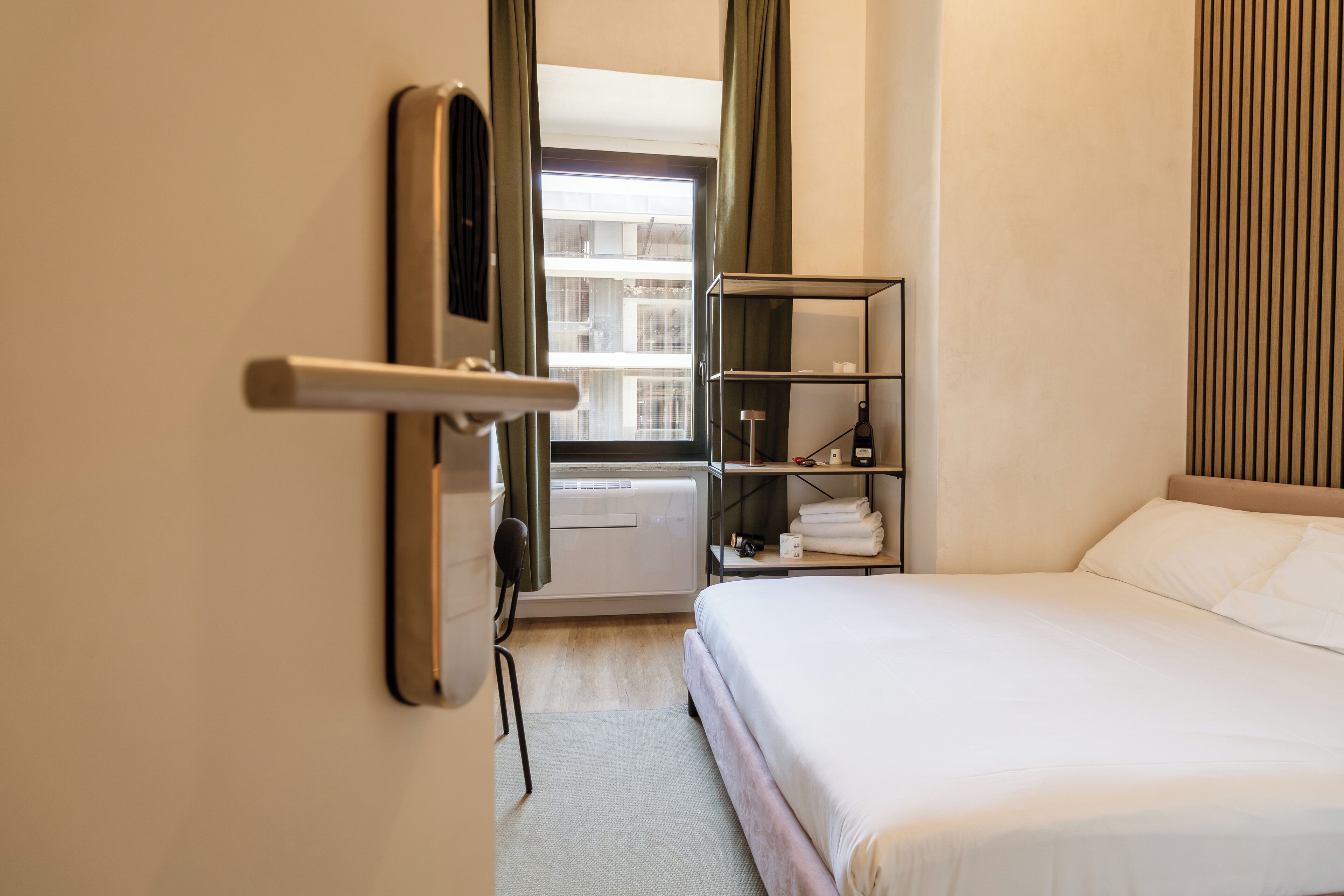 A hotel room with a bed, a shelf with towels, a radiator, and a window with curtains.