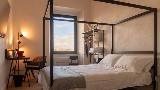 A bedroom with a four-poster bed, a desk with a chair, a small table, and a window with a view of the city. - Cunabu Apartments Giolitti (Rome)