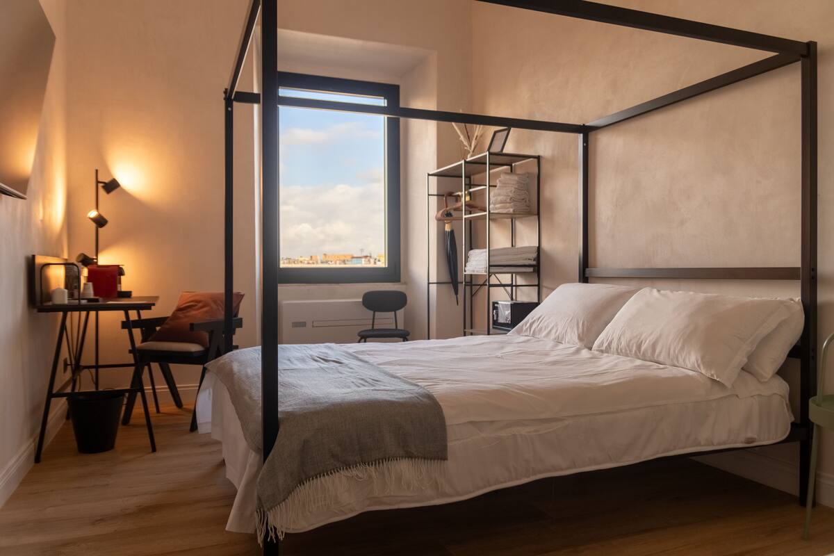 Quarto com cama de dossel, escrivaninha com cadeira, mesinha e janela com vista para a cidade.