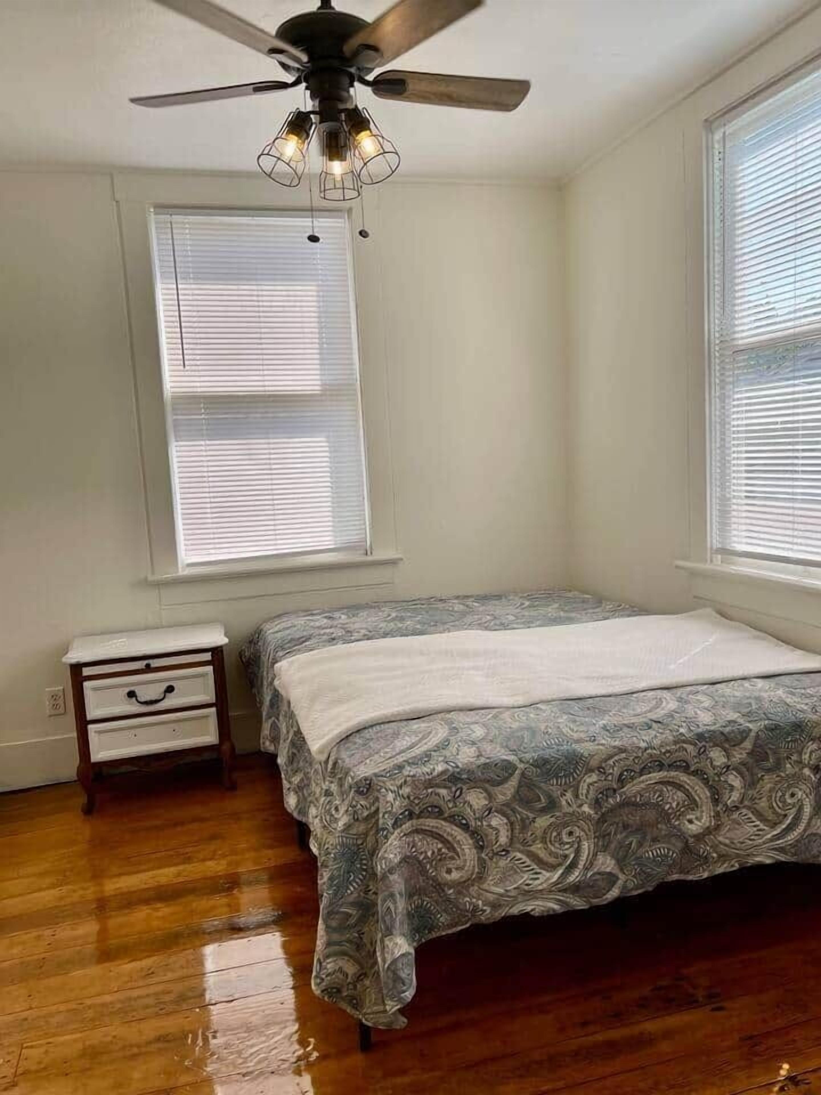 A bedroom with a bed, a nightstand, a ceiling fan, and two windows with blinds.