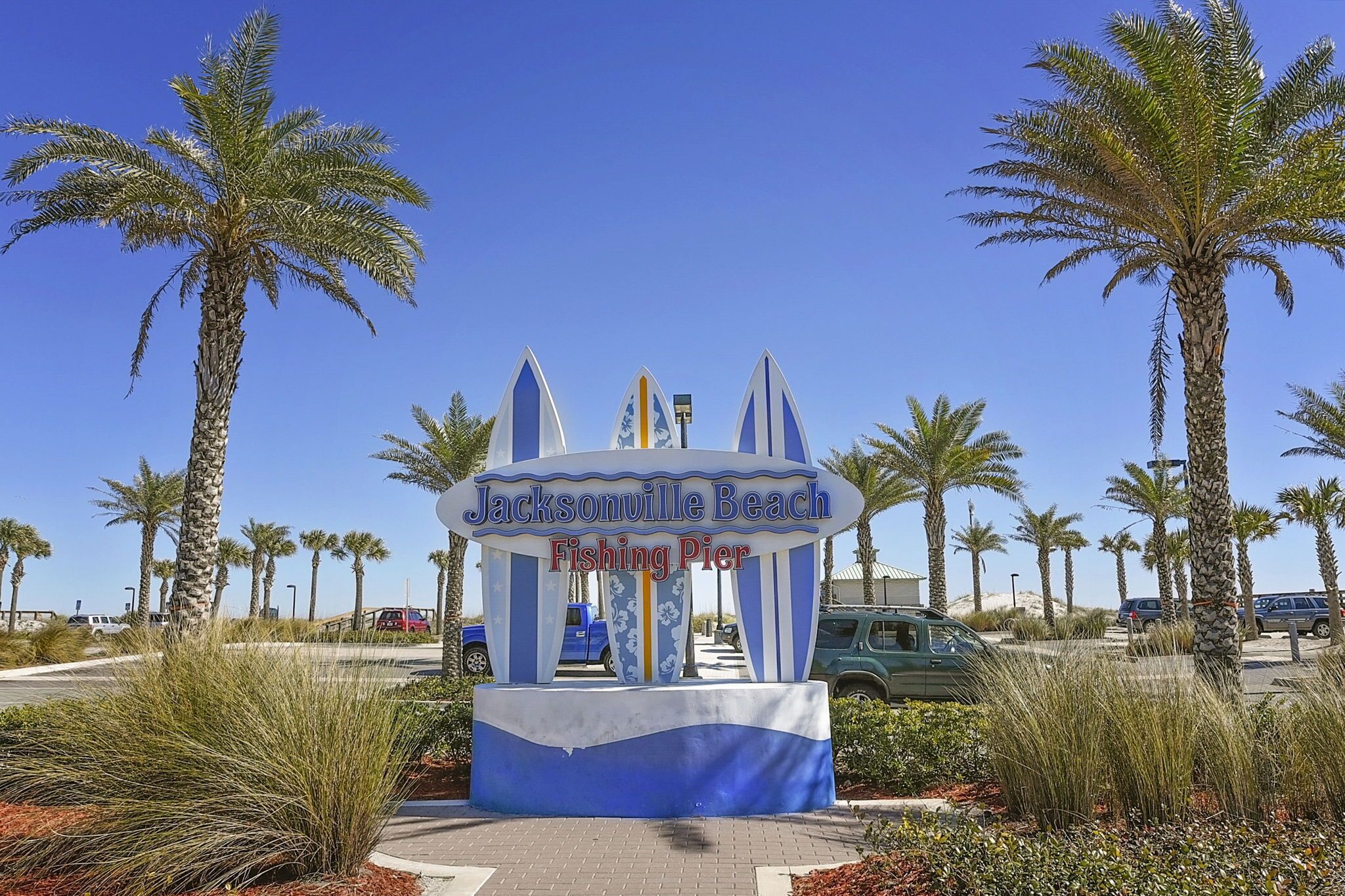 Señal para el muelle de Jacksonville Beach, con palmeras y coches estacionados al fondo.