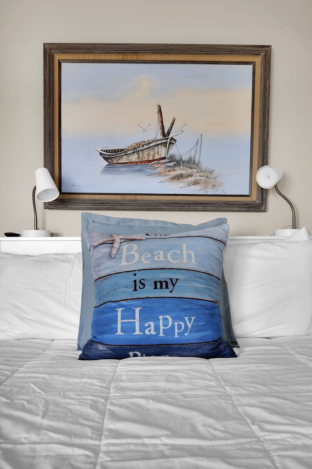A bed with a pillow that reads "Beach is my Happy" and a framed painting of a boat on water.