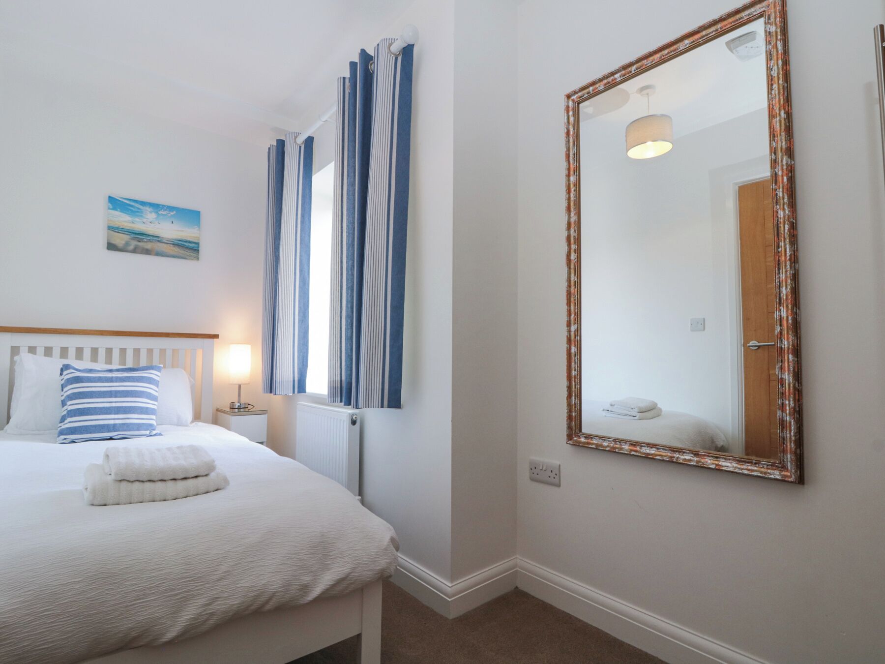 A neatly made bed with white bedding and a blue striped pillow, a large mirror with a decorative frame, and a window with blue curtains.
