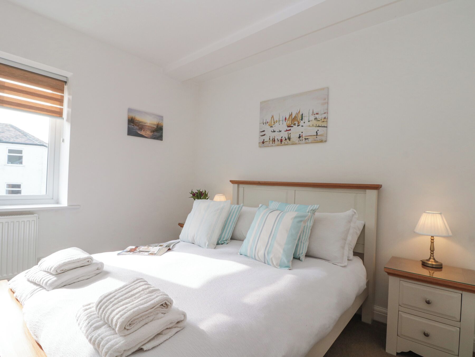 A neatly made bed with white linens and striped pillows, a bedside table with a lamp, and a painting of a sailboat on the wall.