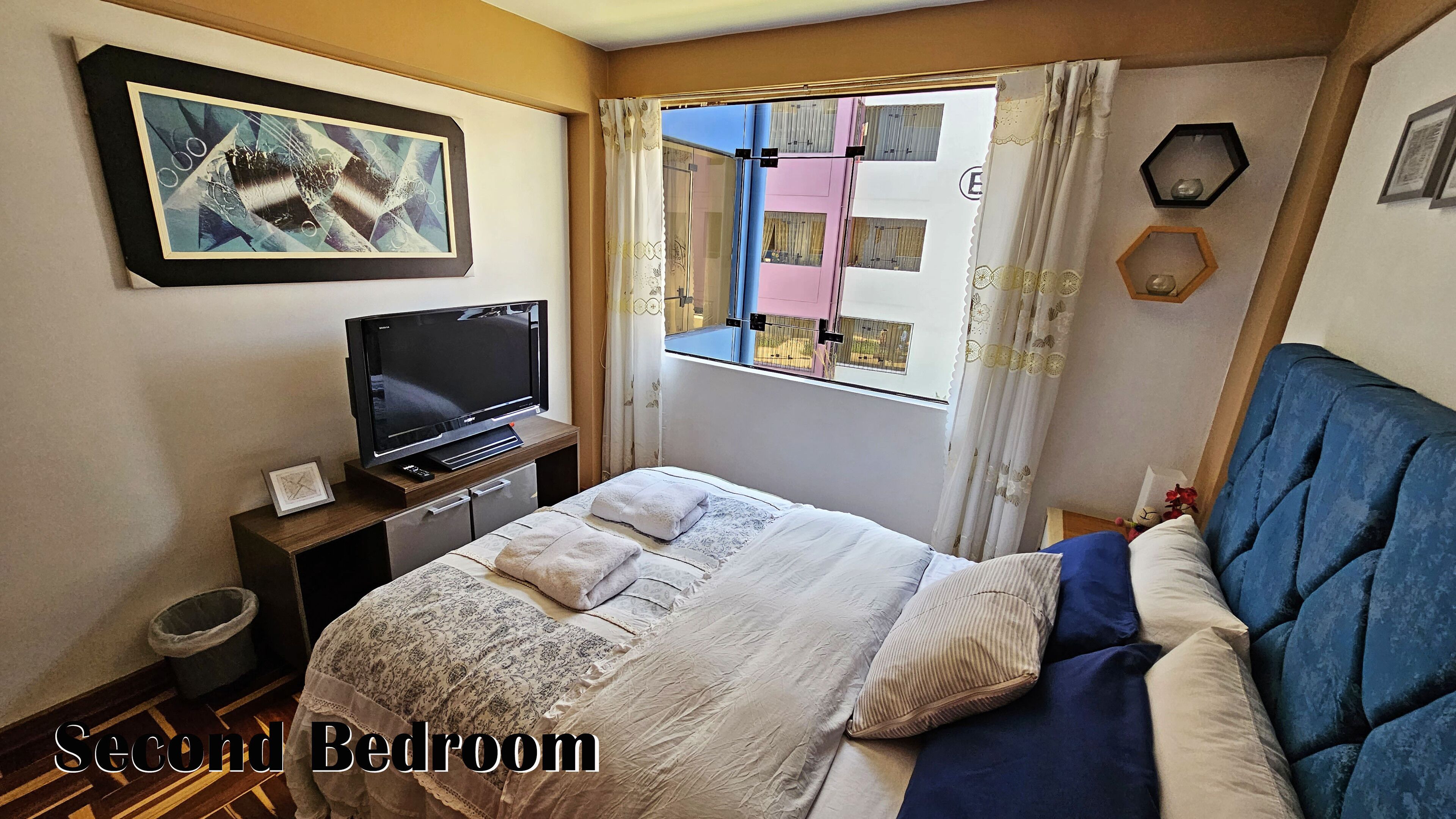 Een slaapkamer met een bed, een televisie, een raam met gordijnen, een foto aan de muur en een prullenbak.