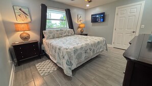 A bedroom with a bed, two bedside tables, a ceiling fan, a TV, a window with curtains, and a door.