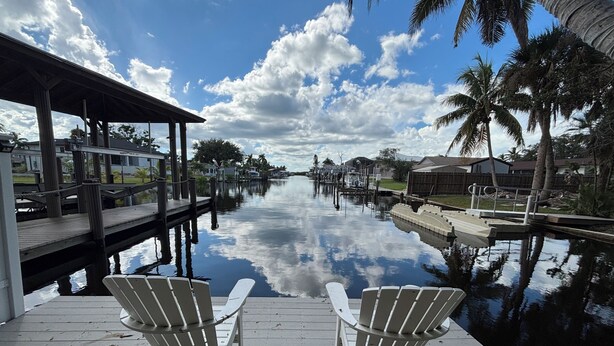 A wooden dock with two Adirondack chairs facing a calm water body, surrounded by palm trees and residential buildings.