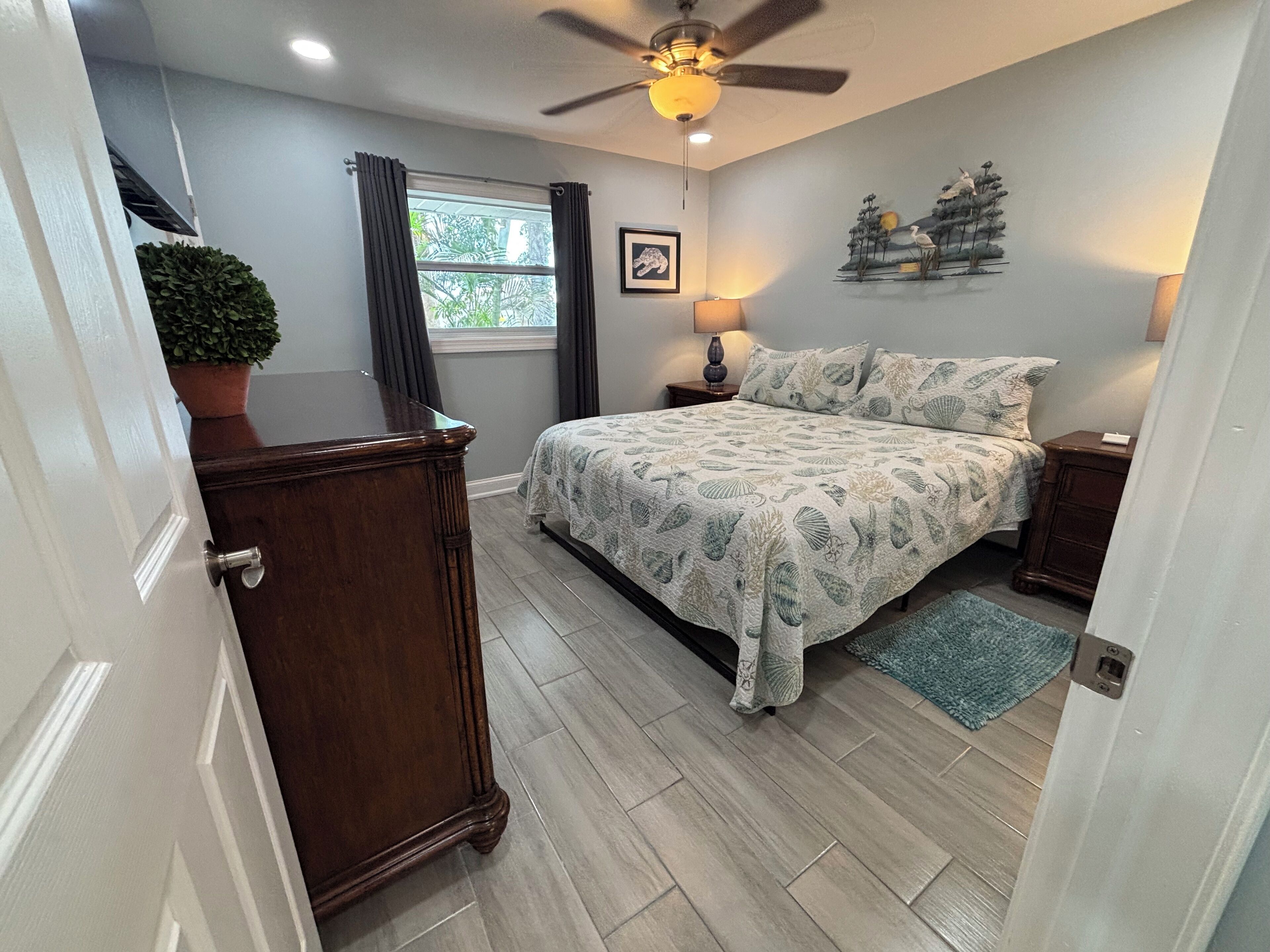 A bedroom with a bed, a nightstand, a lamp, a ceiling fan, a window with curtains, a potted plant, and a wall decoration.