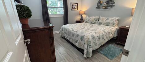 A bedroom with a bed, a nightstand, a lamp, a ceiling fan, a window with curtains, a potted plant, and a wall decoration.
