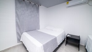 A single bed with a white bedspread in a room with white walls and a white floor. There is a small bedside table with a lamp and a power outlet. An air conditioning unit is mounted on the wall above the bed. - HOTEL SANTA MARTA TROPICAL (Santa Marta)
