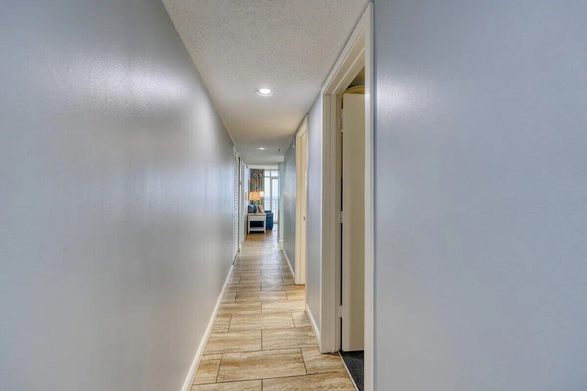 A hallway with light-colored walls and a wooden floor, leading to a room with a window and a door.