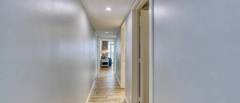 A hallway with light-colored walls and a wooden floor, leading to a room with a window and a door.