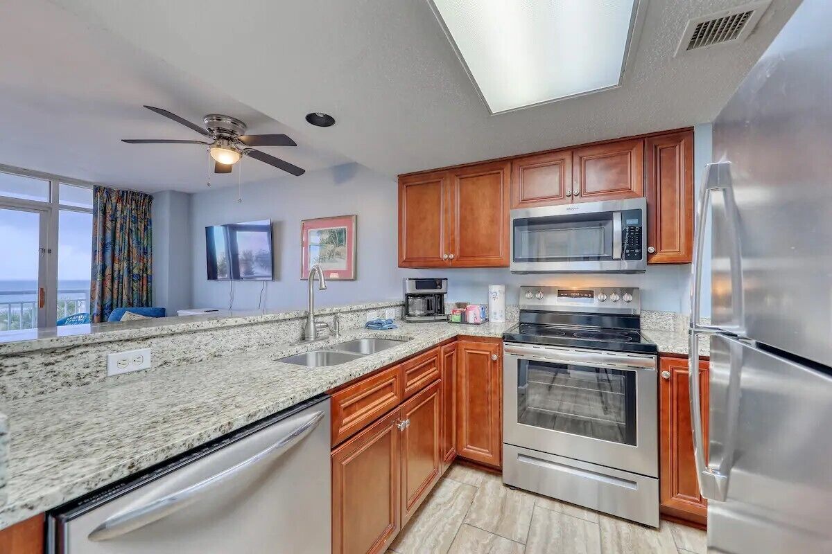 A modern kitchen with granite countertops, stainless steel appliances, wooden cabinets, and a ceiling fan.