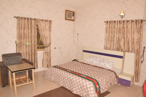 A bedroom with a bed, a chair, a table, and curtains. - See New Hotel (Nsukka)