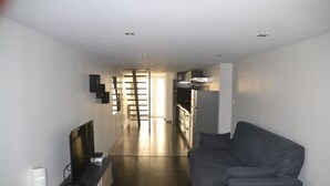 Un appartement moderne doté d’un canapé gris, d’un téléviseur à écran plat, d’une table d’appoint en bois et d’un coin cuisine avec des appareils en acier inoxydable.
