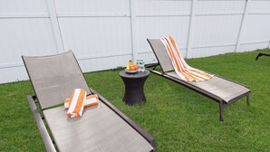 Two outdoor lounge chairs with cushions, a small round table with a bowl of fruit and bottles, and a white fence in the background. - Deluxe Room with Queen Bed and Kitchenette (Clearwater)