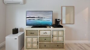 A room with a flat-screen TV mounted on the wall, a small white refrigerator, a wooden cabinet with drawers, and a white air conditioning unit. - Deluxe Room with Queen Bed and Kitchenette (Clearwater)