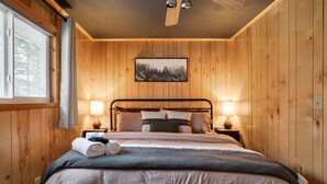 Ett sovrum med träpanel på väggarna, en säng, två sänglampor, en takfläkt och en inramad tavla på väggen.
