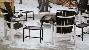 En snötäckt uteplats med Adirondack-stolar och ett bord, en hög med ved och en stuga med en skorsten.