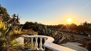 A balcony with a white balustrade overlooking a garden with palm trees and a sunset in the background.