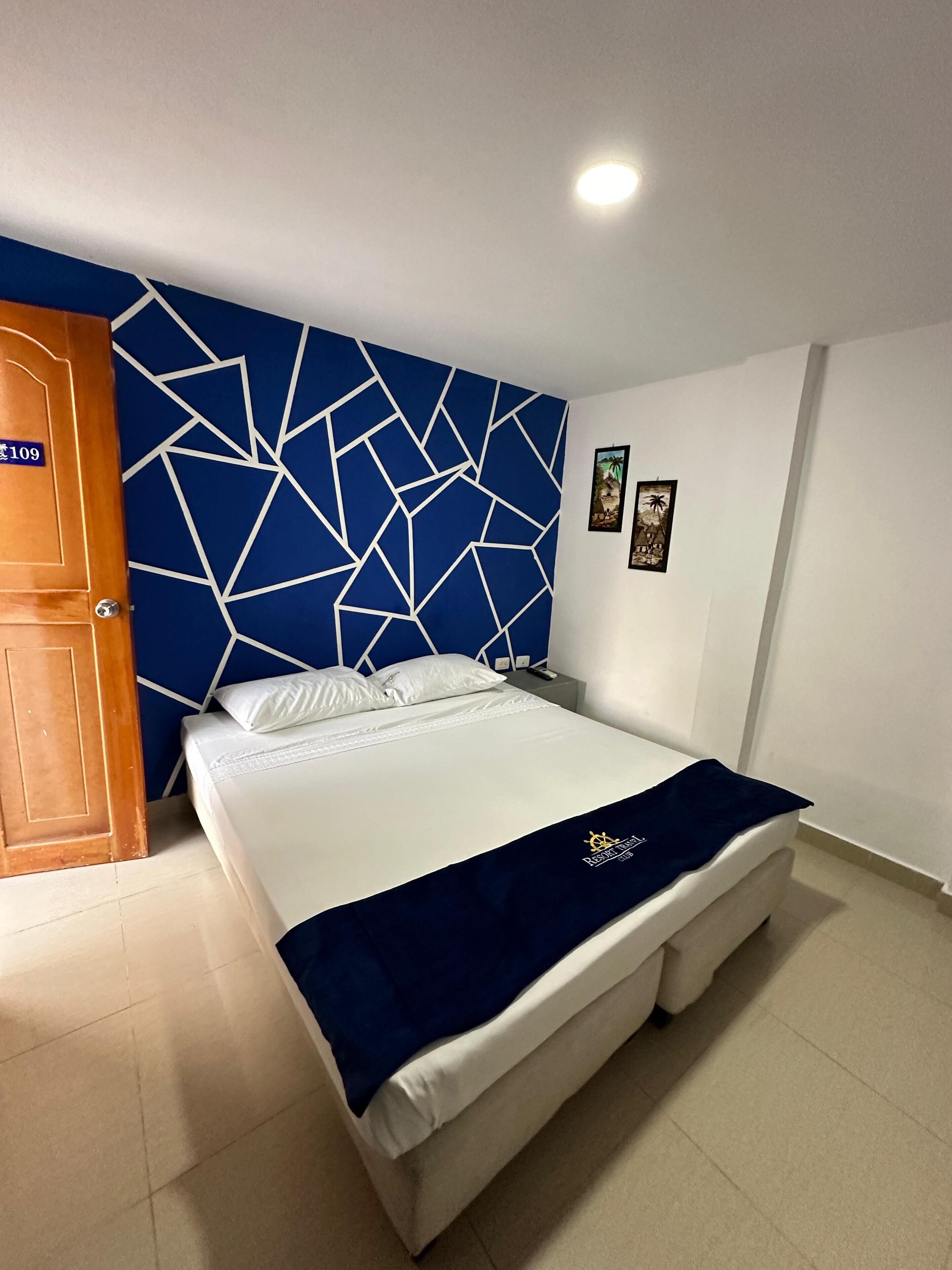 A hotel room with a large bed, a blue geometric-patterned headboard, a white bedside table, and a door with a number sign.