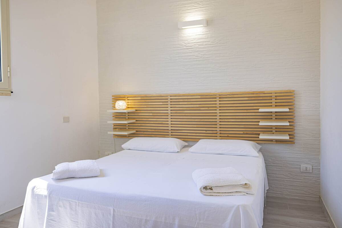Un lit bien fait, avec du linge de lit et des serviettes blanches, une tête de lit en bois et un interrupteur sur le mur.