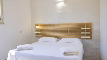 Una cama bien hecha con sábanas y toallas blancas, cabecera de madera y un interruptor de luz en la pared.