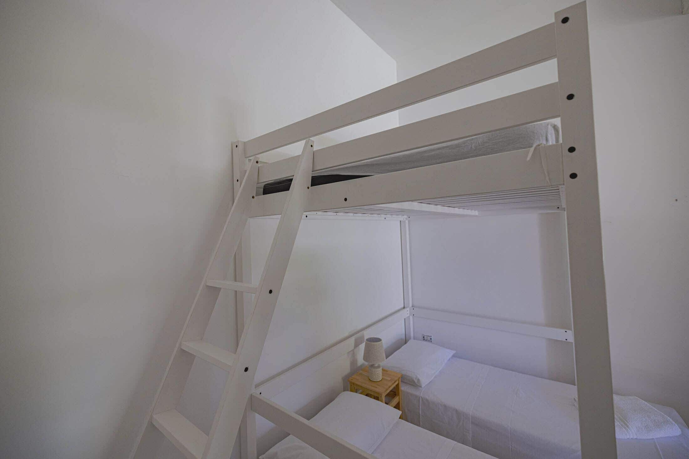 Un lit superposé avec une échelle, une petite table de chevet en bois avec une lampe et un oreiller blanc.