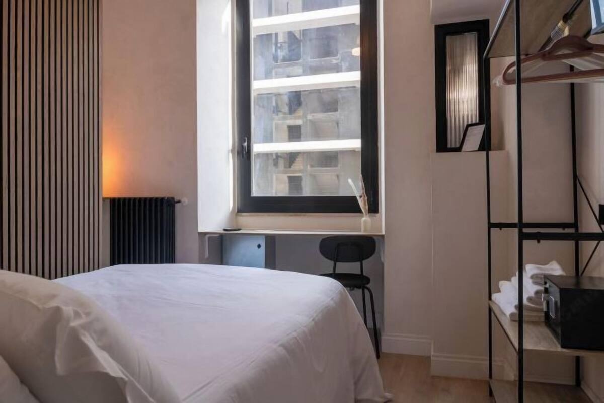 A bedroom with a bed, a window with blinds, a radiator, a chair, and a shelving unit.
