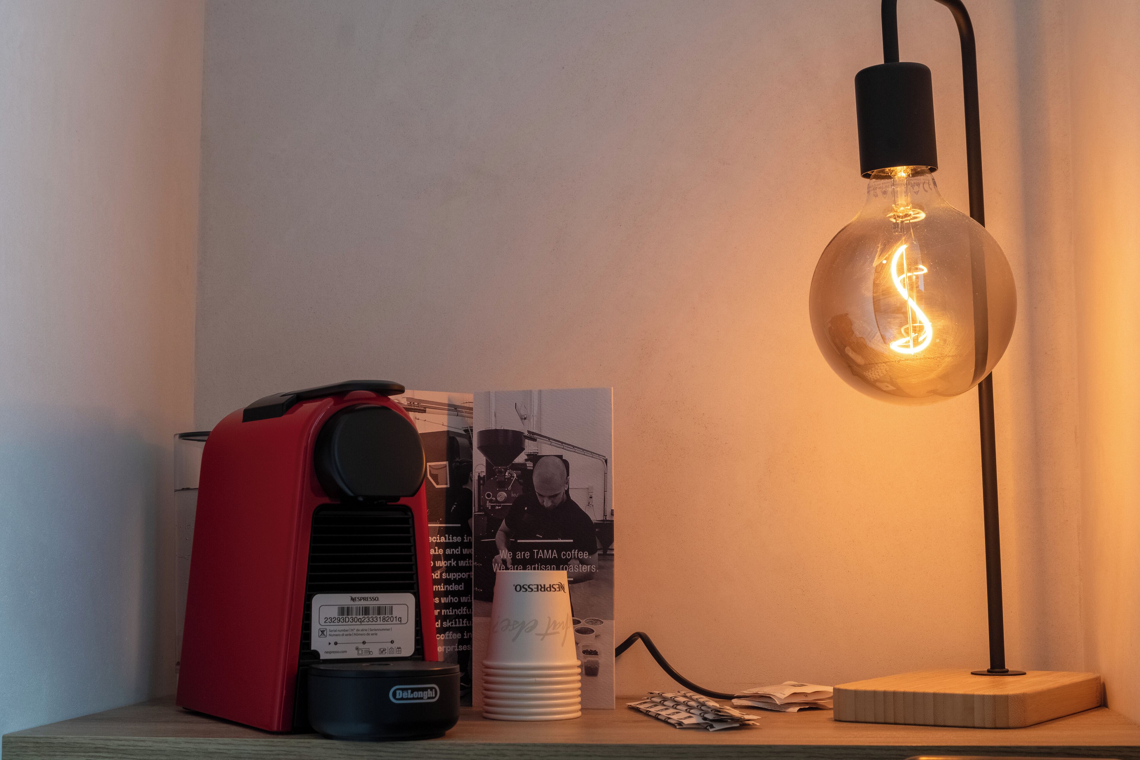 Una cafetera, una pila de vasos desechables, un cartel y una lámpara de mesa sobre una superficie de madera.